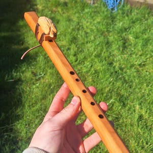 May include: A light brown wooden flute with a smooth, polished finish. The instrument features five circular finger holes along the front and a carved wooden block secured to the top with a dark leather cord, creating a traditional wind instrument.