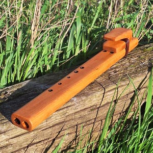 Puede incluir: Una flauta de madera hecha a mano, de color marrón rojizo. El instrumento presenta un bloque rectangular en la parte superior y varios agujeros para los dedos a lo largo del cuerpo. Descansa sobre una superficie de madera envejecida, rodeada de hierba verde. Mide unos 30 cm.