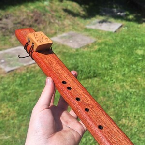 May include: A wooden flute with a reddish-brown finish. The instrument has a decorative wooden block at the top and several finger holes. The flute is held against a backdrop of green grass and foliage.