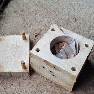 May include: A wooden money box with a circular cavity, containing folded banknotes. The box is square with holes in each corner. A separate wooden lid with dowel pins is next to the box. The wood has a natural, unfinished appearance.