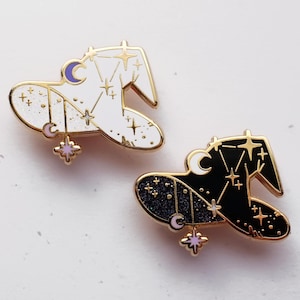May include: Two celestial-themed enamel pins. One is white with gold accents, a crescent moon, and star details. The other is black with gold stars and a crescent moon. Both have a dangling star charm.