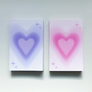May include: Two rectangular notepads with heart designs. One notepad features a purple heart, the other a pink heart, both with a soft, blurred effect. Each notepad has star accents.