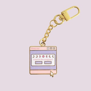May include: A gold-coloured keychain featuring a charm in the shape of a computer window. The window is pink and purple with the text "Sleep.exe" and "Launch the program?" The charm has a gold chain and clasp.