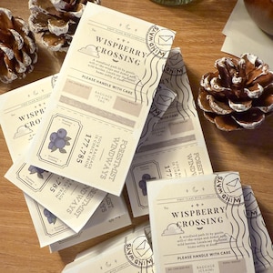 Puede incluir: Una colección de etiquetas de papel color crema con el texto "Wispberry Crossing" e ilustraciones de arándanos y nubes. Las etiquetas tienen una estética vintage y están esparcidas sobre una superficie de madera, junto a piñas.