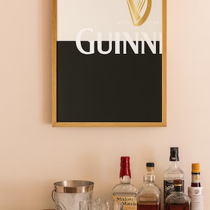 K&ouml;nnte beinhalten: Gerahmtes Guinness-Poster mit goldenem Harfenlogo und Markennamen in Wei&szlig;. Dar&uuml;ber ein Barwagen mit Spirituosenflaschen, einem Eisk&uuml;bel, Gl&auml;sern und einer Holzschale mit Limetten.