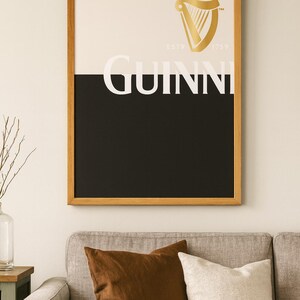 K&ouml;nnte beinhalten: Gerahmtes Guinness-Poster mit goldenem Harfenlogo und dem Wort "GUINNESS" in Wei&szlig;. Das Poster ist in schwarze und wei&szlig;e Abschnitte unterteilt und befindet sich in einem Holzrahmen. Es h&auml;ngt &uuml;ber einem grauen Sofa mit braunen und wei&szlig;en Kissen.