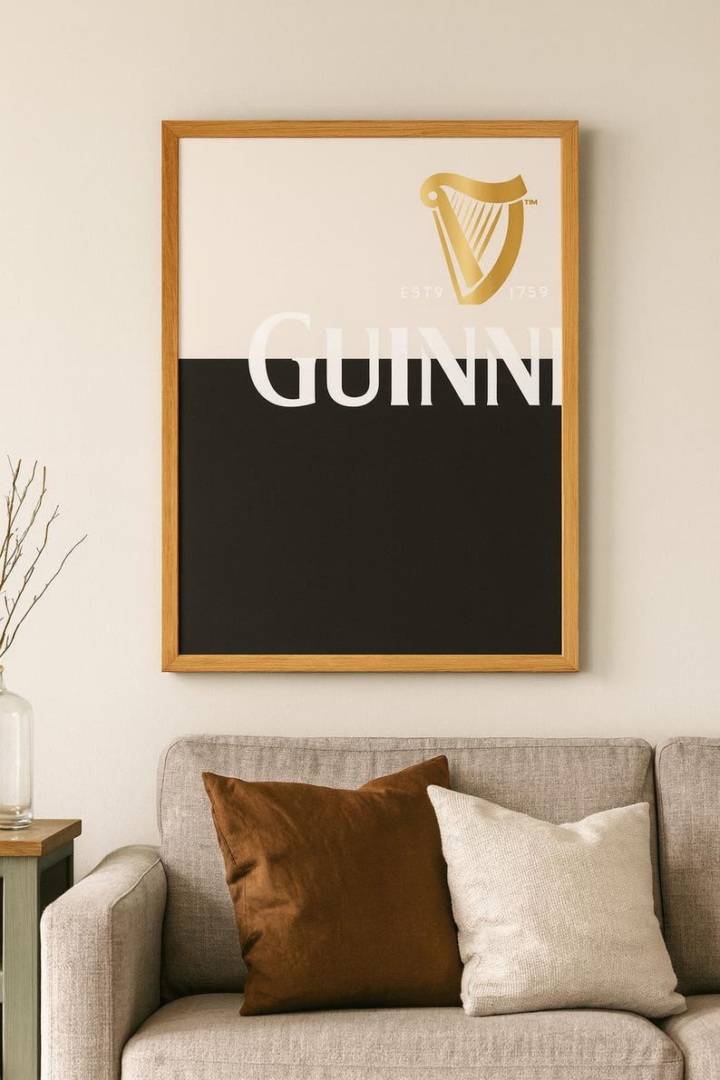 K&ouml;nnte beinhalten: Gerahmtes Guinness-Poster mit goldenem Harfenlogo und dem Wort "GUINNESS" in Wei&szlig;. Das Poster ist in schwarze und wei&szlig;e Abschnitte unterteilt und befindet sich in einem Holzrahmen. Es h&auml;ngt &uuml;ber einem grauen Sofa mit braunen und wei&szlig;en Kissen.