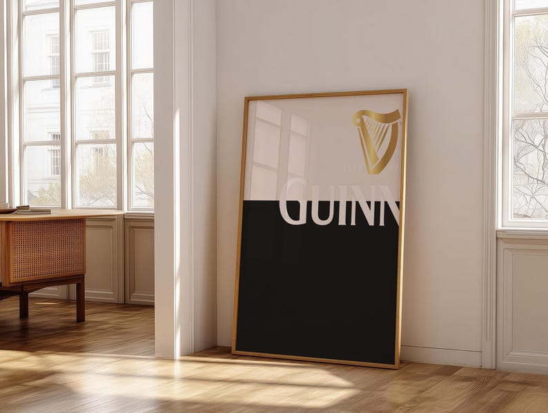 K&ouml;nnte beinhalten: Gerahmtes Guinness-Poster mit goldenem Rahmen. Die obere H&auml;lfte ist wei&szlig;, die untere schwarz, mit dem Wort "GUINNESS" in Wei&szlig;. Ein goldenes Harfenlogo befindet sich in der oberen rechten Ecke. Das Poster lehnt an einer wei&szlig;en Wand.