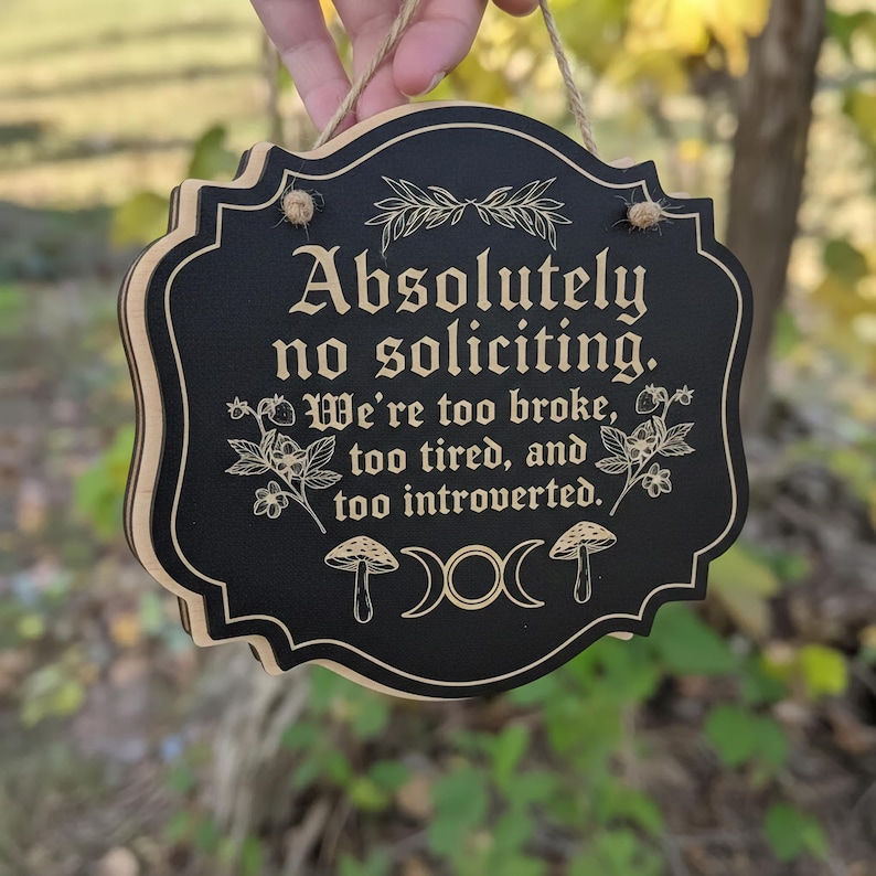 May include: A black wooden sign with the text "Absolutely no soliciting. We're too broke, too tired, and too introverted." The sign has a decorative border and is suspended by a jute rope.