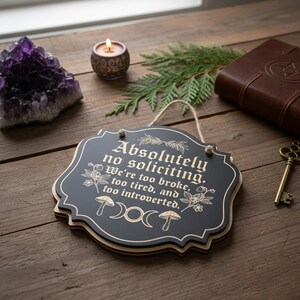 May include: A black wooden sign with the text "Absolutely no soliciting. We're too broke, too tired, and too introverted." The sign has a decorative border and is hanging by a string. Other items include a purple crystal, a candle, and a key.