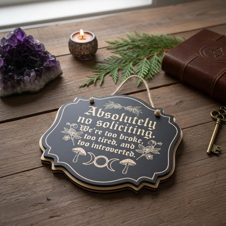 May include: A black wooden sign with the text "Absolutely no soliciting. We're too broke, too tired, and too introverted." The sign has a decorative border and is hanging by a string. Other items include a purple crystal, a candle, and a key.