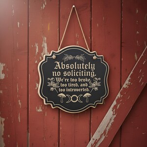 May include: A black wooden sign with a decorative border and the text "Absolutely no soliciting. We're too broke, too tired, and too introverted." The sign is hanging on a red wooden door with a rope.