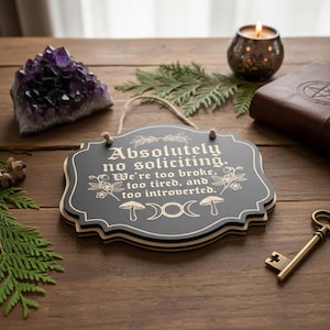 May include: A black wooden sign with the text "Absolutely no soliciting. We're too broke, too tired, and too introverted." The sign has a decorative border and is hanging by a twine string. Other items include a purple crystal, a candle, and a key.