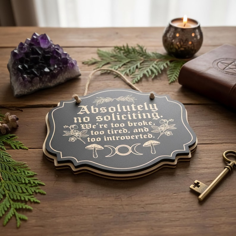 May include: A black wooden sign with the text "Absolutely no soliciting. We're too broke, too tired, and too introverted." The sign has a decorative border and is hanging by a twine string. Other items include a purple crystal, a candle, and a key.