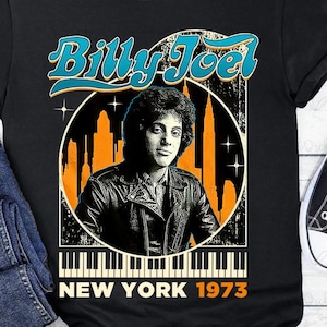 May include: Black t-shirt featuring a graphic of Billy Joel in a leather jacket, set against a New York City skyline. The design includes the text "Billy Joel" in teal and "New York 1973" in white.