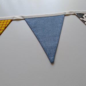 Puede incluir: Una guirnalda de tela con tres banderines triangulares unidos a una cinta de color crema. El bander&iacute;n izquierdo tiene un patr&oacute;n de cuadr&iacute;cula amarillo y rojo, el central es de mezclilla azul y el derecho tiene un estampado de vaca.