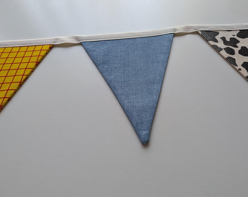 Puede incluir: Una guirnalda de tela con tres banderines triangulares unidos a una cinta de color crema. El bander&iacute;n izquierdo tiene un patr&oacute;n de cuadr&iacute;cula amarillo y rojo, el central es de mezclilla azul y el derecho tiene un estampado de vaca.
