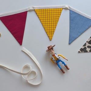 Puede incluir: Una guirnalda de tela con banderines triangulares en rojo s&oacute;lido, amarillo con cuadr&iacute;cula roja, azul s&oacute;lido y estampado de manchas de vaca en blanco y negro. Una insignia de estrella de sheriff de madera y una figura de vaquero est&aacute;n debajo.