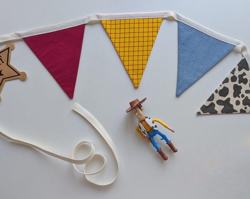 Puede incluir: Una guirnalda de tela con banderines triangulares en rojo s&oacute;lido, amarillo con cuadr&iacute;cula roja, azul s&oacute;lido y estampado de manchas de vaca en blanco y negro. Una insignia de estrella de sheriff de madera y una figura de vaquero est&aacute;n debajo.