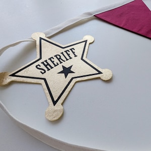 Puede incluir: Una insignia de estrella de sheriff de tela dorada unida a una cinta blanca. La estrella presenta un contorno negro, la palabra SHERIFF impresa en letras may&uacute;sculas negras y un peque&ntilde;o icono de estrella negra. Se ve un bander&iacute;n triangular rojo oscuro.