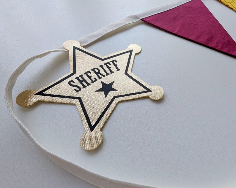 Puede incluir: Una insignia de estrella de sheriff de tela dorada unida a una cinta blanca. La estrella presenta un contorno negro, la palabra SHERIFF impresa en letras may&uacute;sculas negras y un peque&ntilde;o icono de estrella negra. Se ve un bander&iacute;n triangular rojo oscuro.