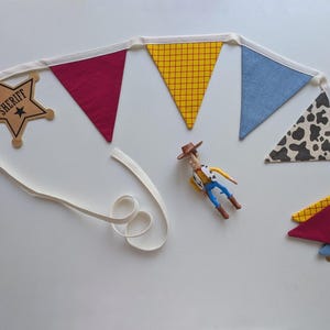 Puede incluir: Una guirnalda de tela con una estrella de sheriff de madera, banderines triangulares rojos, amarillos a cuadros, azul claro y con estampado de vaca unidos a una cinta blanca, junto a una peque&ntilde;a figura de vaquero de pl&aacute;stico.