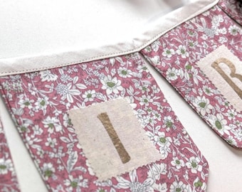 Puede incluir: Una guirnalda de tela con un estampado floral rosa de flores blancas y verdes. Cada banderín triangular está unido a un borde de tela color crema. Un parche cuadrado color crema con una letra I dorada metálica está centrado en un banderín.