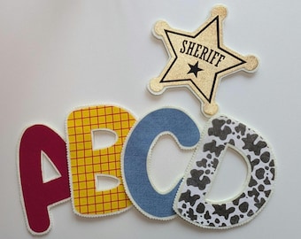 Puede incluir: Un conjunto de cuatro letras de tela que forman ABCD, con una A granate sólida, una B con patrón de cuadrícula amarillo, una C con textura de mezclilla azul y una D con estampado de vaca en blanco y negro, junto a una insignia de estrella dorada con el texto SHERIFF.