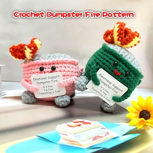 Puede incluir: Dos figuras de ganchillo hechas a mano con forma de cubo de basura en llamas, una rosa y otra verde, cada una con un cartel que dice "Emotional Support Dumpster Fire. It's fine, I'm fine. Everything is fine!". Las figuras tienen caras sonrientes y adornos en forma de llama.