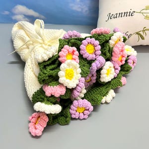 Op de afbeelding: Een crèmekleurige gehaakte tas, vastgebonden met een lint, overvol met een levendig arrangement van gehaakte bloemen. De bloemen hebben roze, witte en paarse bloemblaadjes met gele centra en groene bladeren, wat een vrolijke, handgemaakte esthetiek creëert.