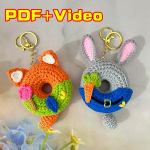 Könnte beinhalten: Zwei handgefertigte Häkel-Schlüsselanhänger. Einer ist ein orangefarbener Fuchs mit weißen Akzenten, einer rosa Blume und einem grünen Blatt. Der andere ist ein graues Kaninchen mit rosa Innenohren, einer blauen Latzhose und einer Karotte. Beide haben goldene Schlüsselringe. Der Text "PDF+Video" steht oben.
