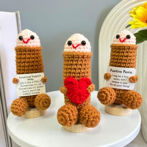 Puede incluir: Tres figuras hechas a mano con ganchillo, con cuerpos marrones, cabezas beige y caras sonrientes. Una sostiene un corazón rojo, otra un letrero que dice "Emotional Support Willy", y la tercera un letrero que dice "Positive Penis". Cada figura está sobre una pequeña base de madera.