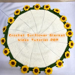 Op de afbeelding: Een ronde, crèmekleurige gehaakte deken met een groene rand en gele en bruine zonnebloemaccenten. De tekst "Crochet Sunflower Blanket Video Tutorial PDF" is oranje weergegeven. De deken is handgemaakt.