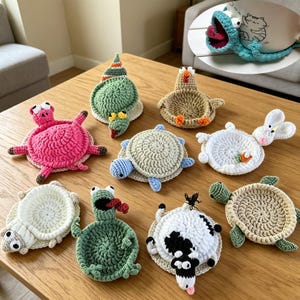 May include: A collection of handmade crochet coasters in various animal shapes, including a pig, turtle, snail, chicken, frog, cow, and rabbit. The coasters are in shades of pink, green, beige, and white, and are arranged on a wooden table.