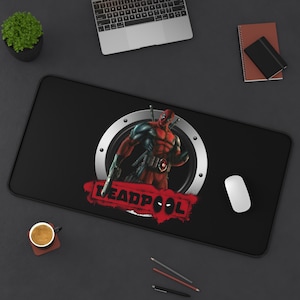 May include: Black rectangular desk mat featuring a Deadpool graphic. The graphic shows Deadpool in a red and black suit, holding a gun, with the word "DEADPOOL" in red. A white computer mouse is on the right.