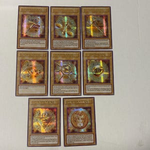Puede incluir: Un conjunto de nueve cartas coleccionables con detalles dorados. Cada carta presenta un diseño único con texto y símbolos hebreos, incluyendo un ojo, una balanza y un ankh. Las cartas están dispuestas en una cuadrícula.