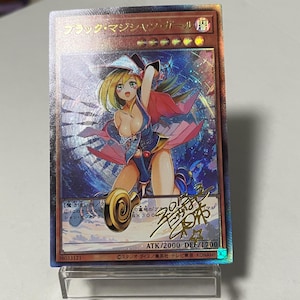Puede incluir: Carta coleccionable holográfica con una ilustración de estilo anime de un personaje femenino con cabello rubio, vestida con un atuendo azul oscuro y rojo. La carta tiene texto japonés y se muestra en un soporte transparente. ATK 2000, DEF 1700.