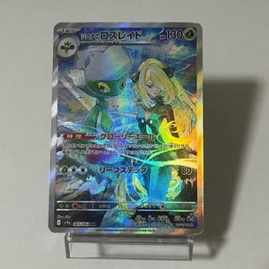 May include: A holographic trading card featuring a green and white floral creature and a blonde woman in a teal dress. The card has Japanese text and the number 130 in the upper right corner. The card is displayed on a clear plastic stand.