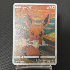 May include: A collectible trading card featuring the brown Eevee character with a surprised expression, set against a vibrant, swirling orange and yellow background. The card displays Japanese text, the name Eevee, and an HP of 60 at the top right.