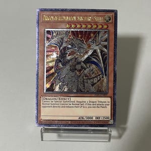 Puede incluir: Carta coleccionable con una ilustración de dragón y detalles en lámina plateada. La carta dice "DRAGON OF ILLUMINATION, SANCTUARY'S SUMMON". La carta se muestra en un soporte transparente.