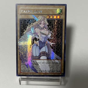 May include: A holographic trading card with the text "HARPIE LADY." The card shows a winged woman in a white and blue outfit. The card has a gold border and includes text such as "YGLD-EN012" and "ATK/1300 DEF/1400." The card is on display.