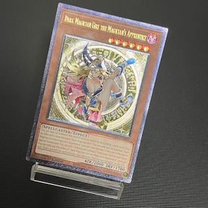 May include: A gold-coloured trading card featuring the character "Dark Magician Girl the Magician's Apprentice." The card has a detailed illustration of the character in a blue and pink outfit. The card text includes "SPELLCASTER/EFFECT" and other game details.