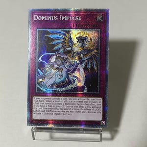 Könnte beinhalten: Holografische Sammelkarte mit dem Text "DOMINUS IMPULSE" oben. Die Karte zeigt eine detaillierte Illustration einer geflügelten Figur mit blauen und goldenen Akzenten. Die Karte wird auf einem transparenten Ständer präsentiert.