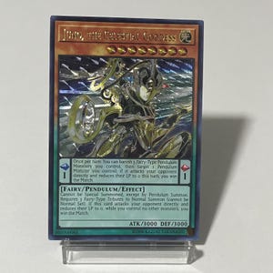 Puede incluir: Una carta de juego que presenta a un personaje femenino con cabello negro y una corona dorada. La carta se titula "JUNO, LA DIOSA CELESTIAL" y tiene un fondo azul con un efecto holográfico. El texto de la carta dice: "Una vez por turno: Puedes desterrar 3 Monstruos Pendulum de Tipo Hada que controles, luego selecciona 1 Monstruo Pendulum que controles; si este ataca directamente a tu oponente y reduce sus LP a 0 este turno, ganas el Duelo."