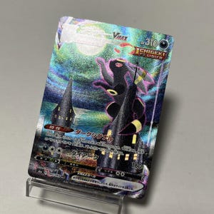 Puede incluir: Una tarjeta coleccionable holográfica con un personaje Umbreon oscuro y acentos morados, sobre un cielo nocturno con dos torres oscuras. La tarjeta tiene texto japonés y una designación VMAX, con 310 HP.