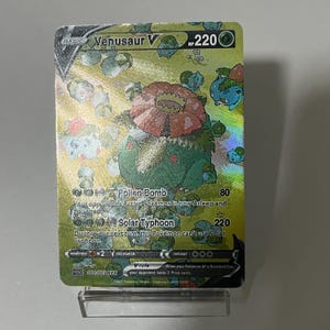 May include: A holographic Venusaur V Pokemon card with a green and red Venusaur illustration. The card has a gold background and the text "Venusaur V" and "HP 220". The card also has two attacks: "Pollen Bomb" and "Solar Typhoon".