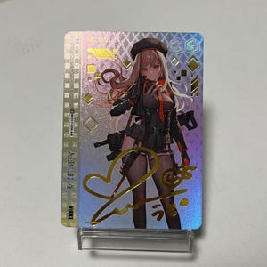 Puede incluir: Tarjeta coleccionable holográfica con un personaje de estilo anime en atuendo negro y naranja, sosteniendo un arma. La tarjeta tiene detalles dorados, texto japonés y la palabra "NIKKE". La tarjeta se muestra en un soporte transparente.