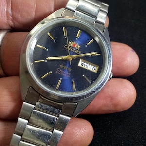 May include: Silver Orient watch with a dark blue face and gold hands and markers. The watch face displays the day and date. The watch has a silver metal band. The watch face has the word "Orient" in gold lettering.