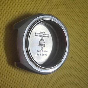 May include: A silver-toned watch case with a circular interior. The watch case has the text "LULETTE PLAQUE OR O BO LUCROUS FOND ACIER WOXYDABLE" and "FIA SUISSE SWISS MADE" printed on the interior. The numbers "166 0174" and "366 0833" are also visible.