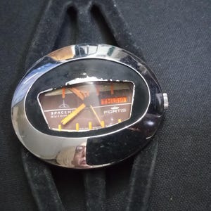 May include: A vintage Fortis Spacematic automatic watch with a black face and silver casing. The watch displays the day and date. The watch has a black strap and is set against a black background.